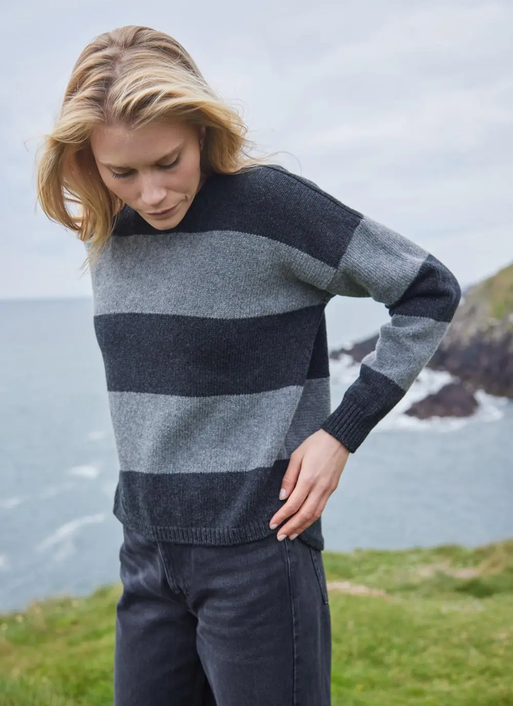 blonde woman looking down standing by Irish cliffs on a windy day wearing a grey stripe sweater with hands on hips