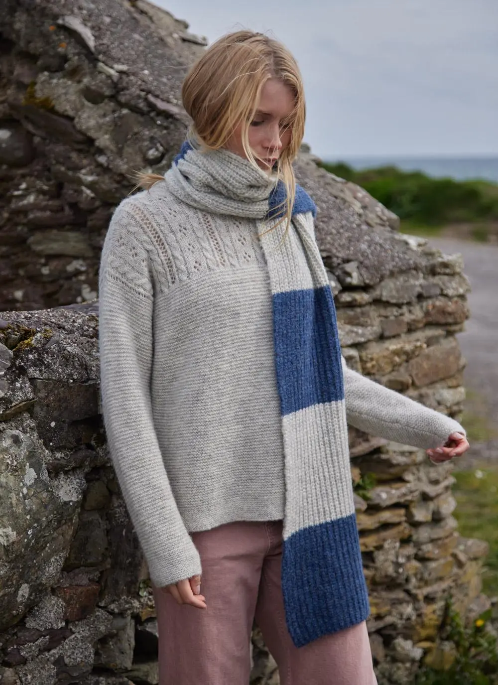blonde woman standing near rocks wearing a large grey and blue knit scarf 