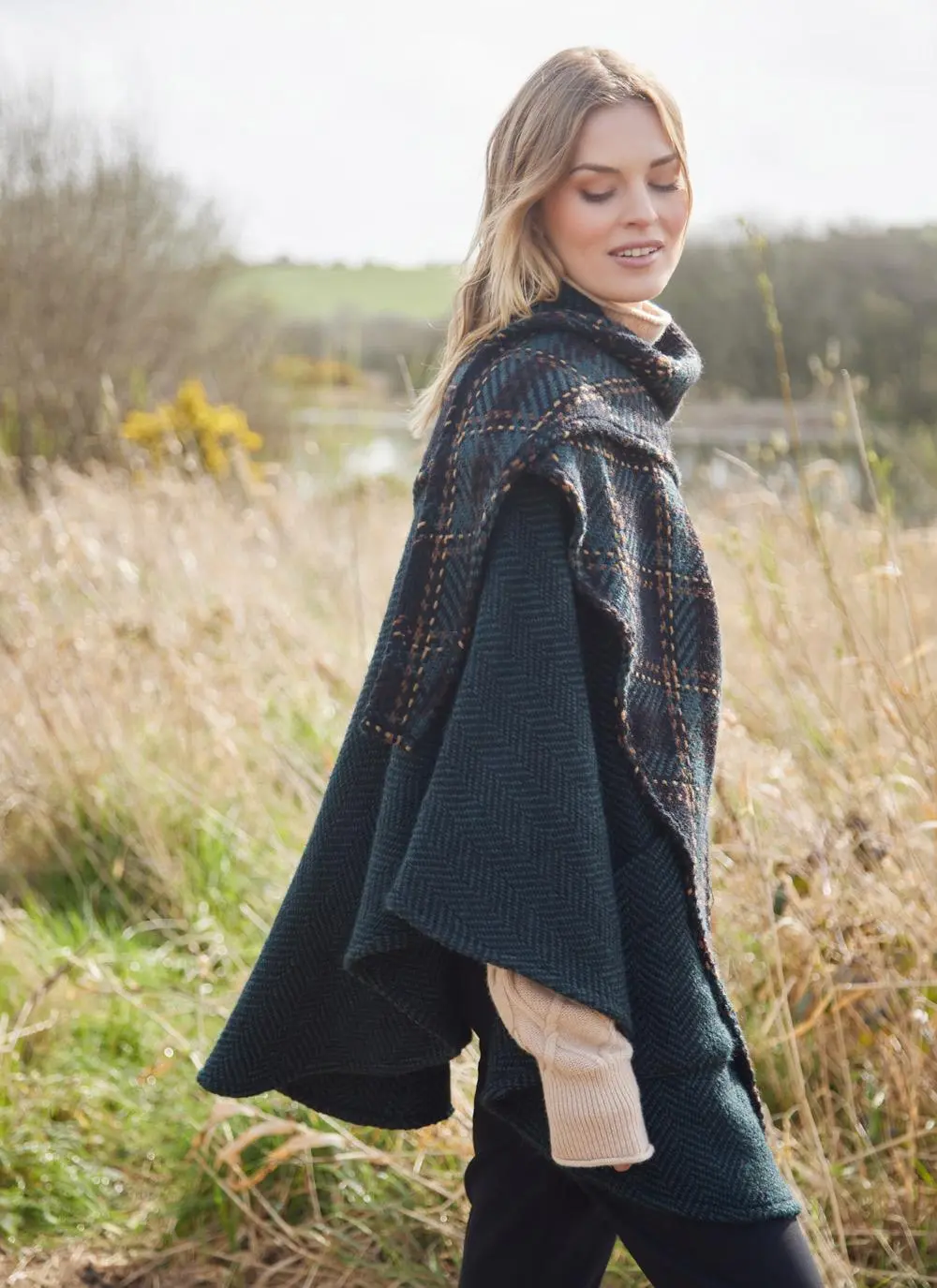 Side angle of blonde woman in grassy Irish countryside wearing dark green herringbone cape.