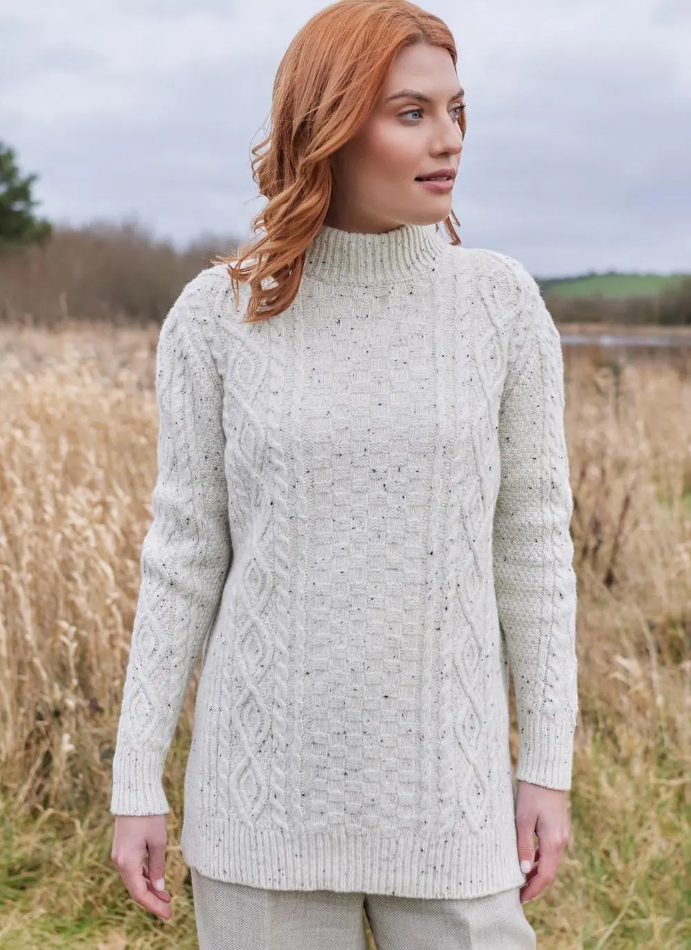 Red haired woman in field wearing ecru colored Aran turtle neck sweater looking to right