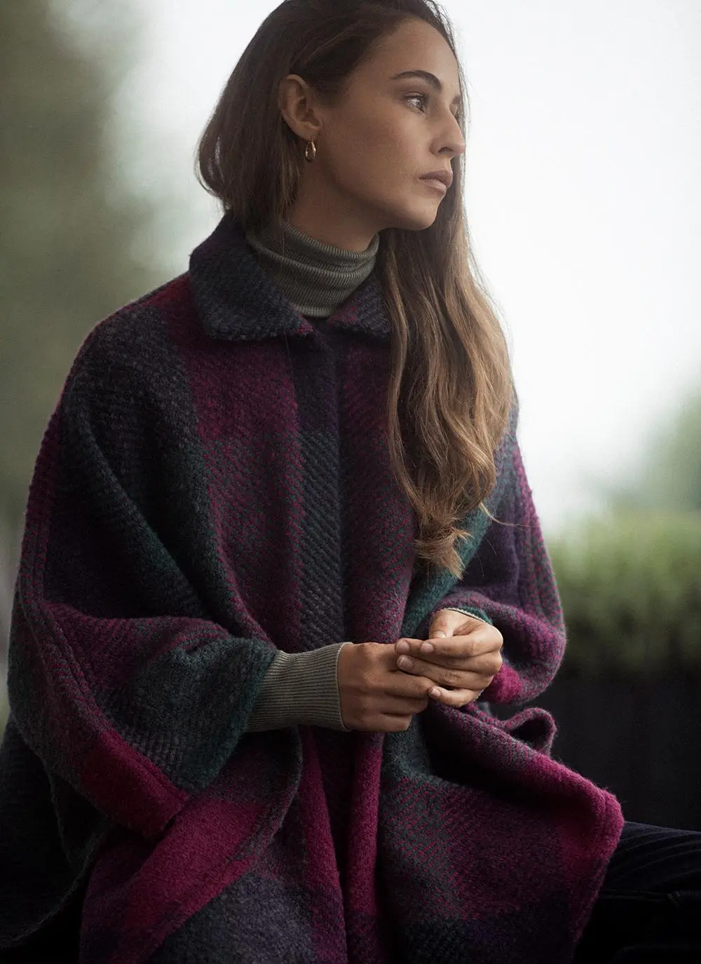 Outdoor shot of woman wearing wool purple toned cape with hands clasped looking to the right.