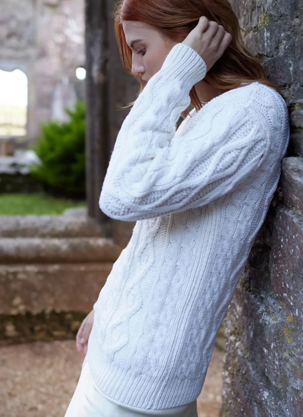 side shot of red haired woman leaning against wall in stony passageway wearing a white aran sweater