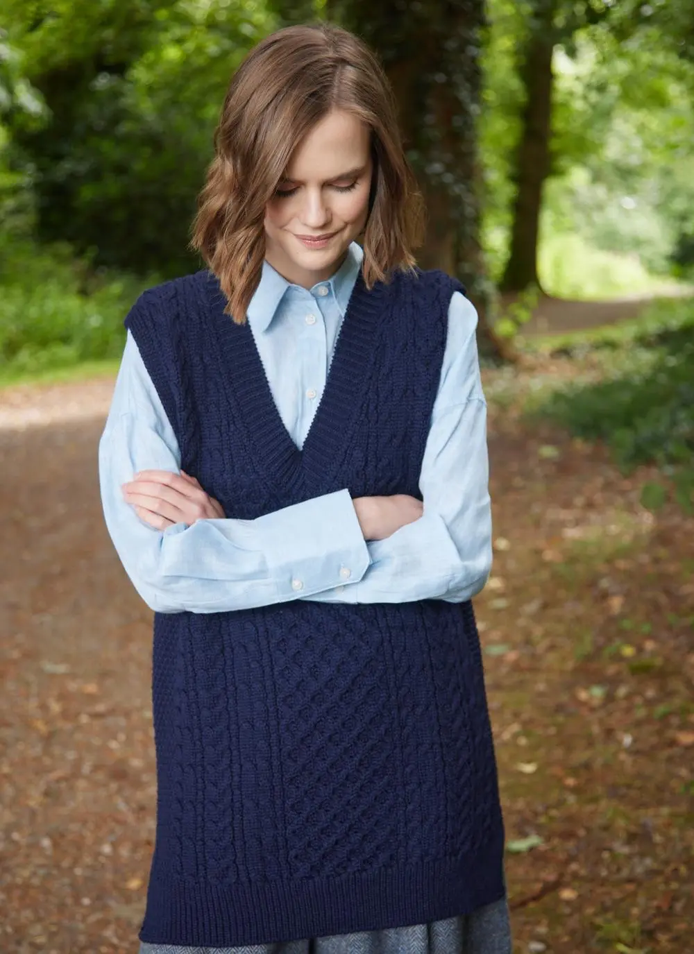 brunette woman standing in forest wearing navy v-neck aran vest with arms folded