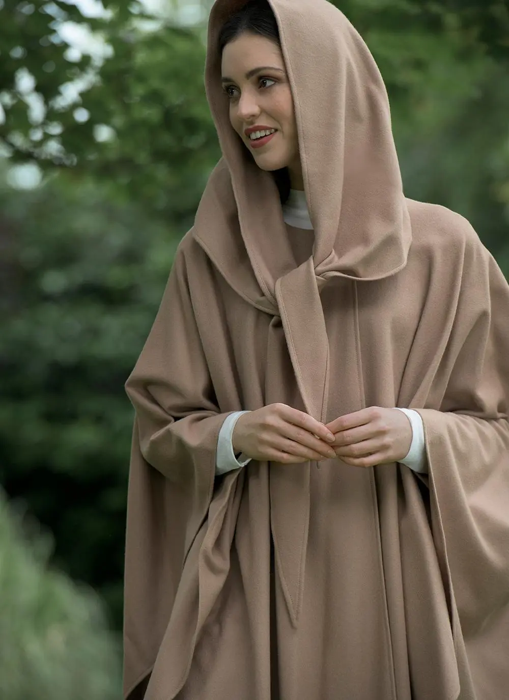 Brunette woman standing outside wearing camel cape with hands clasped.